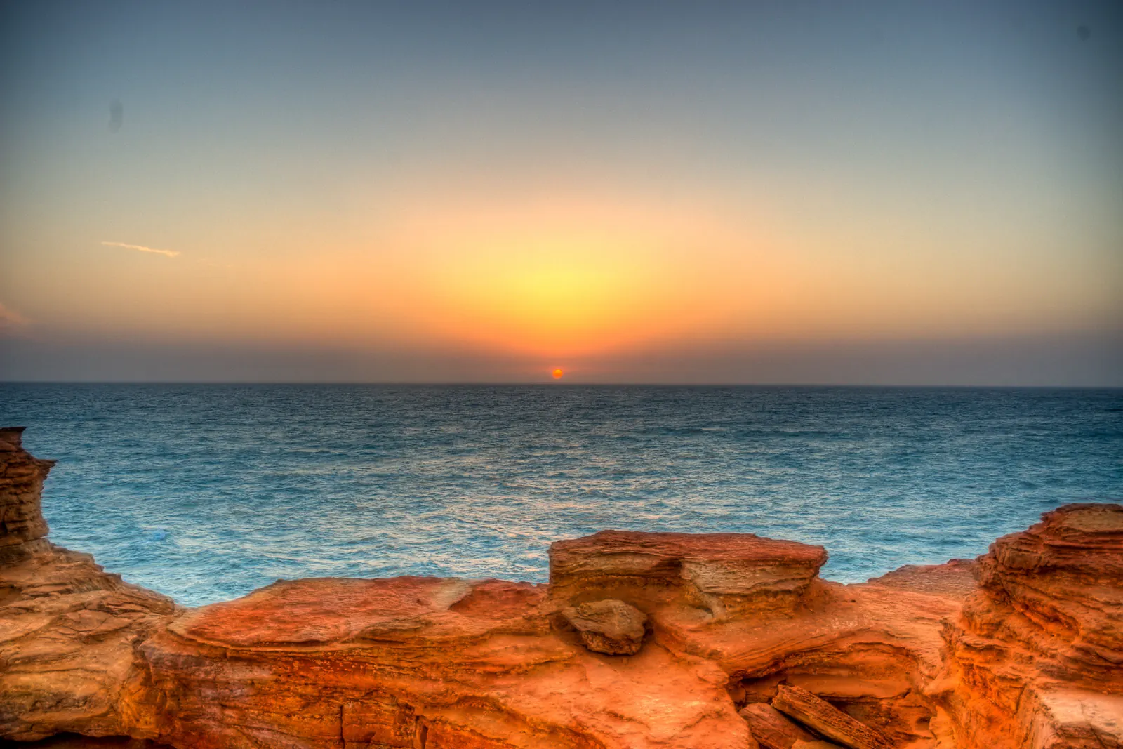 Slunce zapadající do tyrkysového moře za červenými pískovcovými skalami na Gantheaume Point, Broome